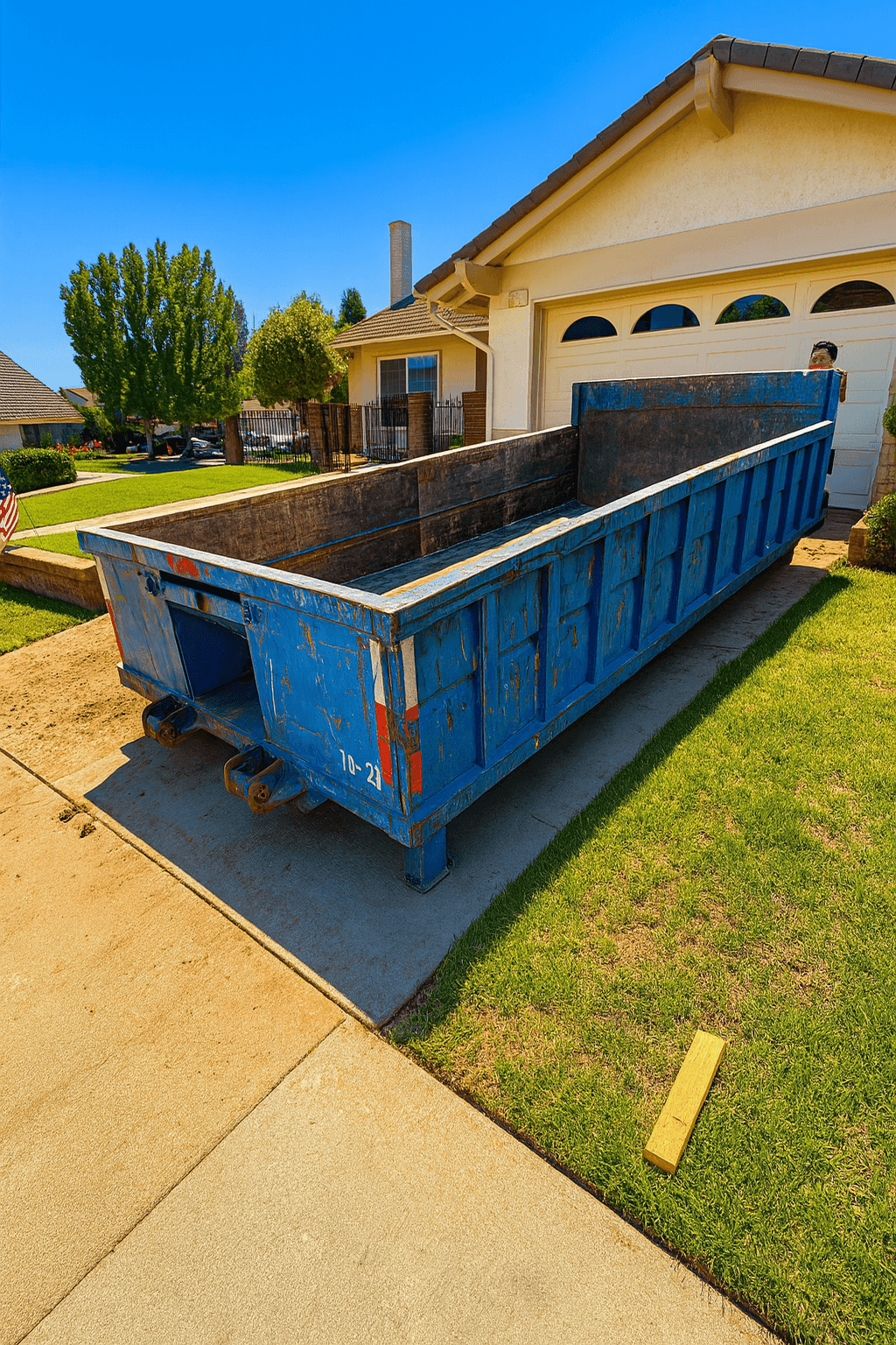 Large blue dumpster placed on a driveway in front of a suburban house under a clear blue sky.