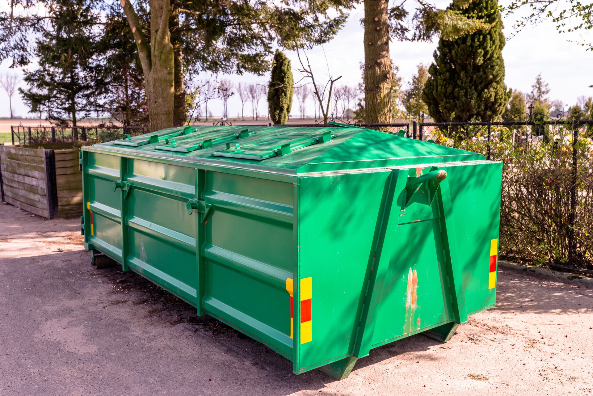A Large, Metal, Green Garbage Container And Municipal Waste, Standing On A Dirt Road Near The Fence
